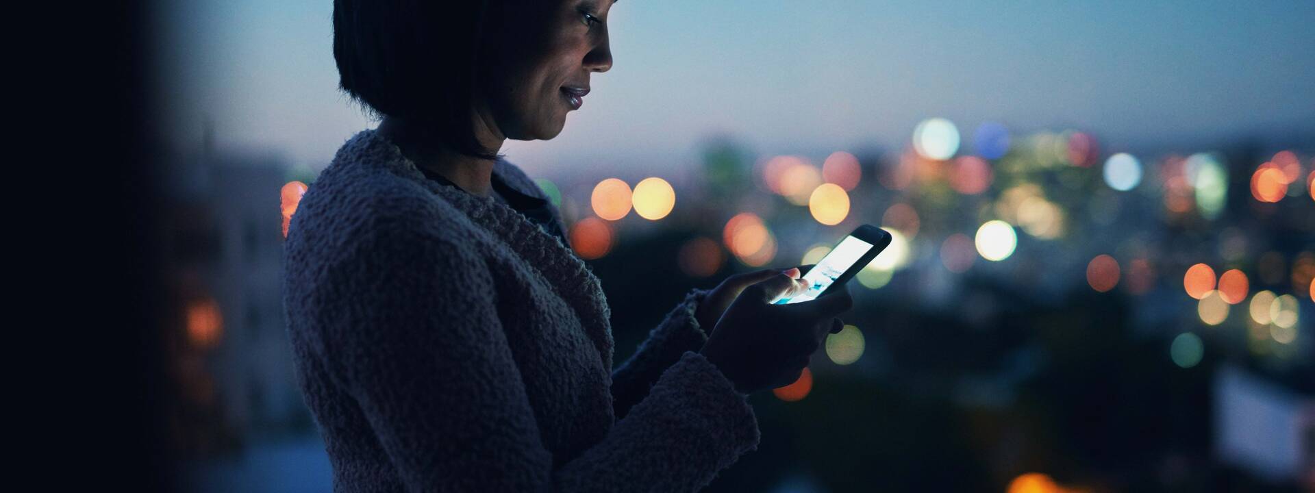 Eine junge Frau telefoniert abends auf einem Balkon. Stadtlichter im Hintergrund